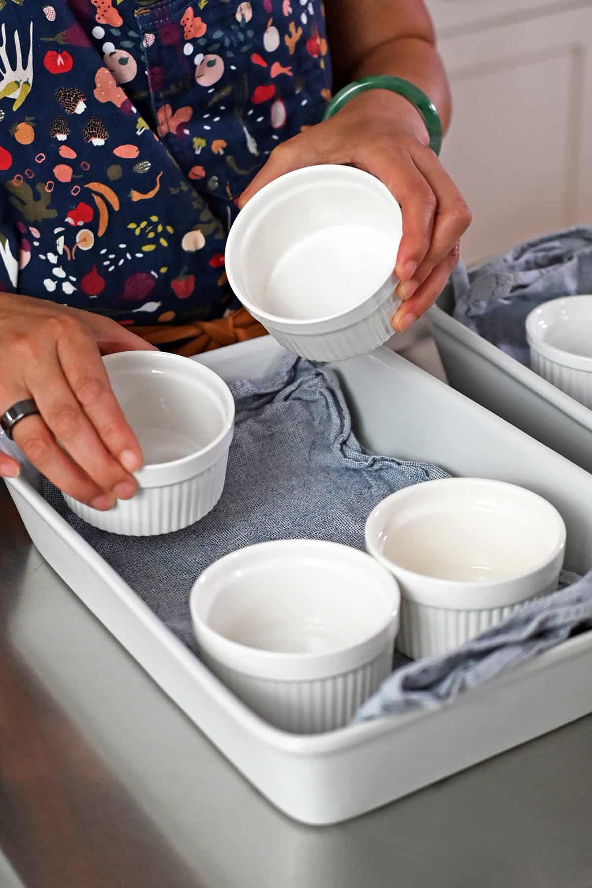 A hand is placing a ramekin into a casserole dish lined with a blue cloth.