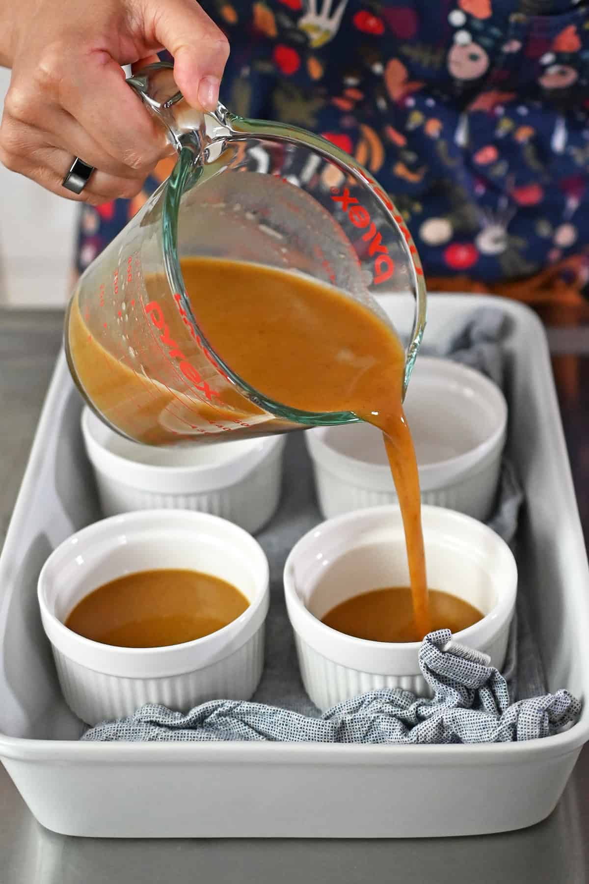 Pouring the pumpkin custard mixture into white ramekins in a casserole dish lined with a blue and white towel.