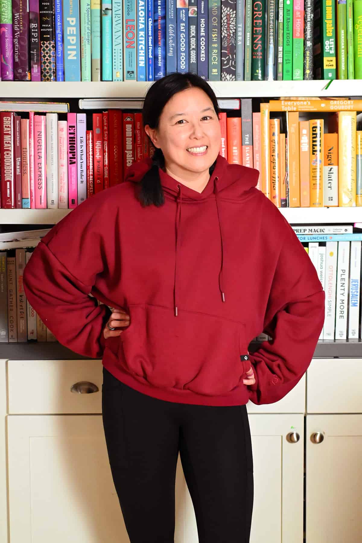 A smiling Asian woman in a red Popflex cloud hoodie and black leggings in front of a bookcase filled with colorful cookbooks.