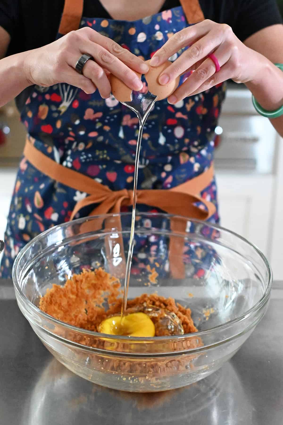 A person in a blue apron is cracking an egg into a bowl filled with coconut sugar, melted coconut oil, and vanilla extract.
