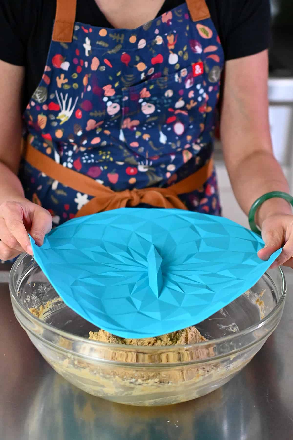 Covering a bowl filled with raw snickerdoodle dough with a blue silicone lid before chilling it in the fridge.