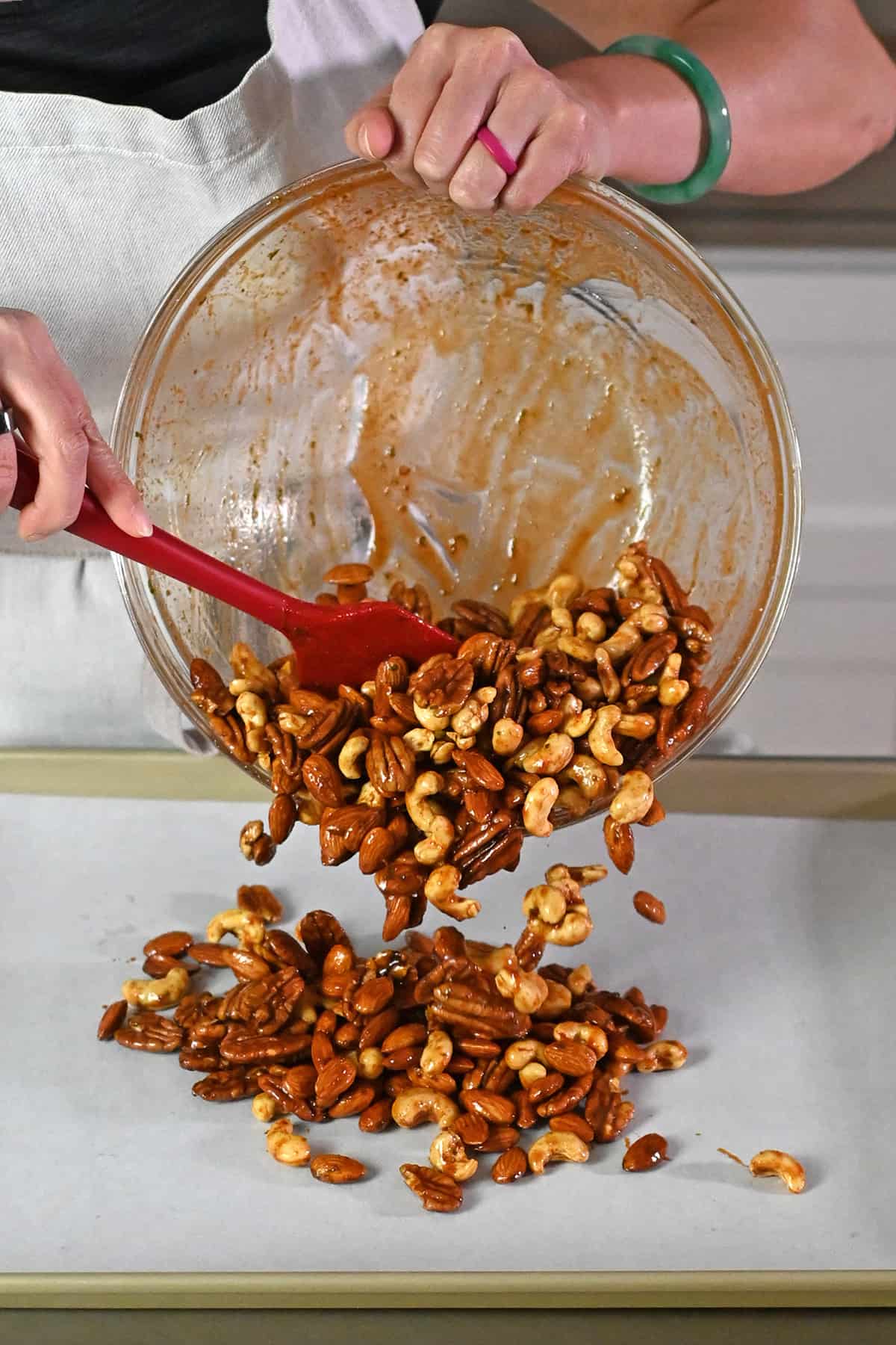 Transferring the seasoned nuts from a large glass mixing bowl to a parchment lined rimmed baking sheet.