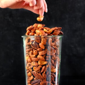 A hand grabbing a cashew from a tall glass jar filled with paleo spiced nuts.