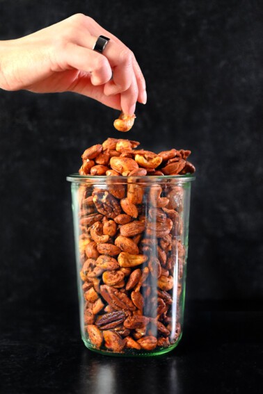 A hand grabbing a cashew from a tall glass jar filled with paleo spiced nuts.