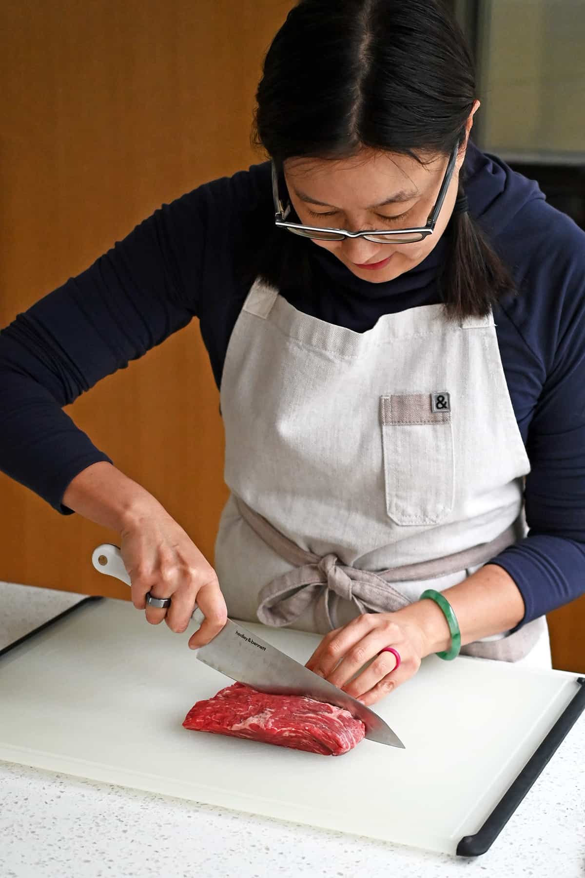An Asian woman is slicing a raw piece of flank steakin half with the grain.