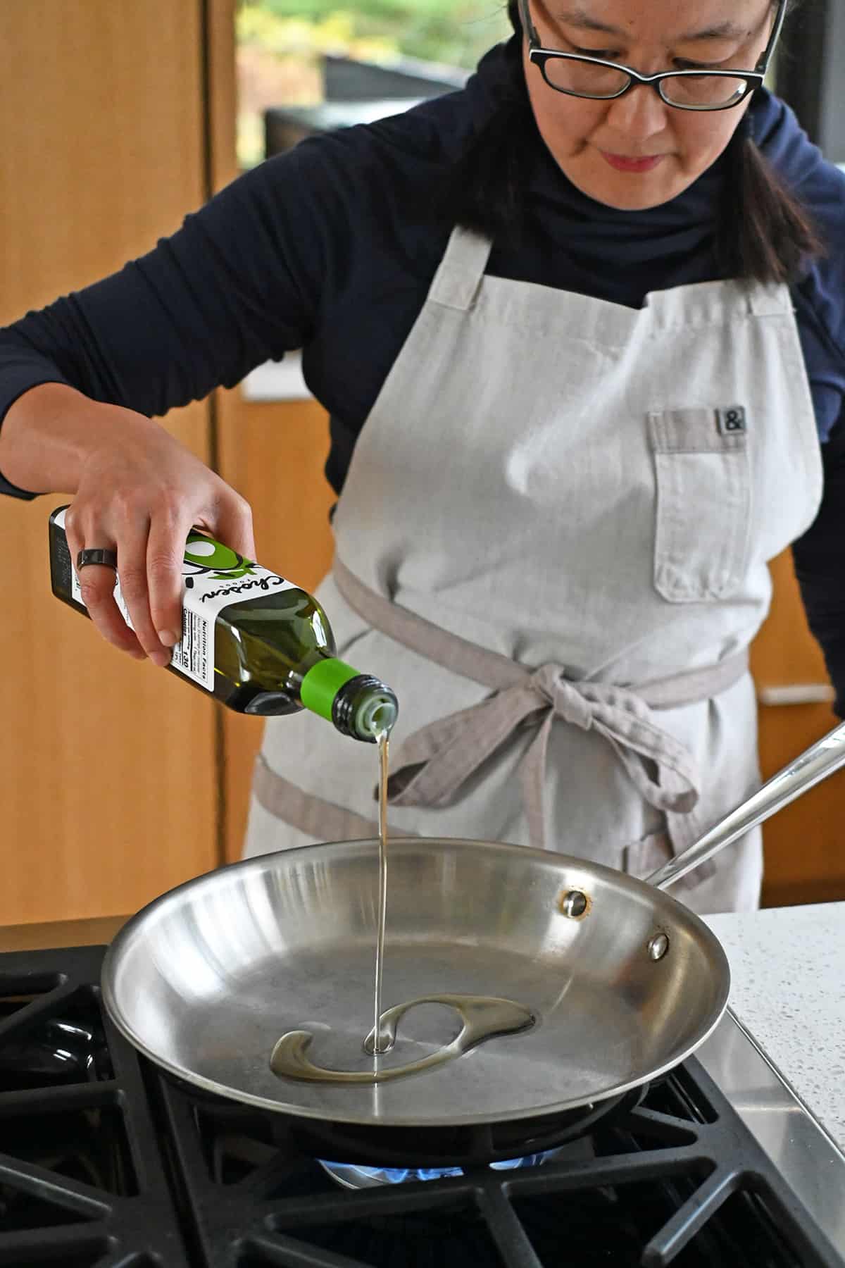 An Asian woman adds avocado oil into a hot large stainless steel skillet.