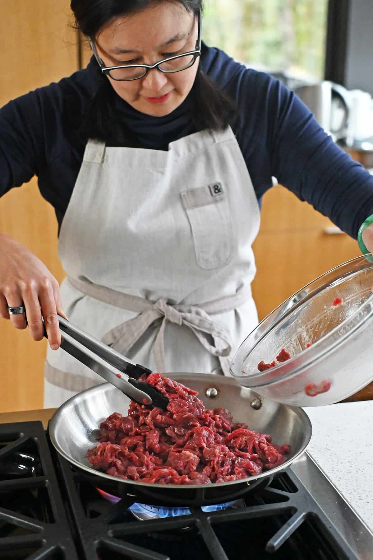 An Asian woman adds marinated sliced flank steak into a hot skillet to make paleo pepper steak.