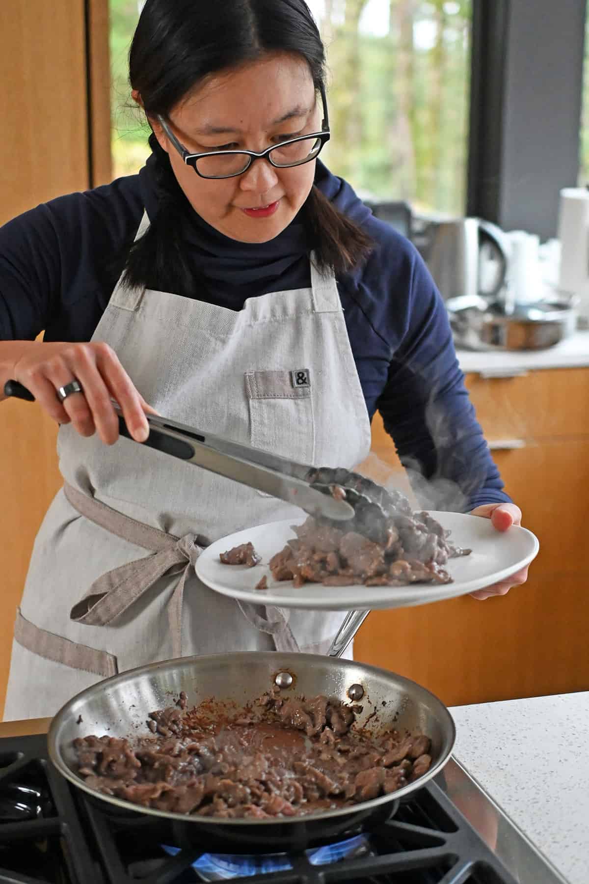 An Asian woman transfers cooked sliced flank steak from a skillet to a plate with a pair of tongs.