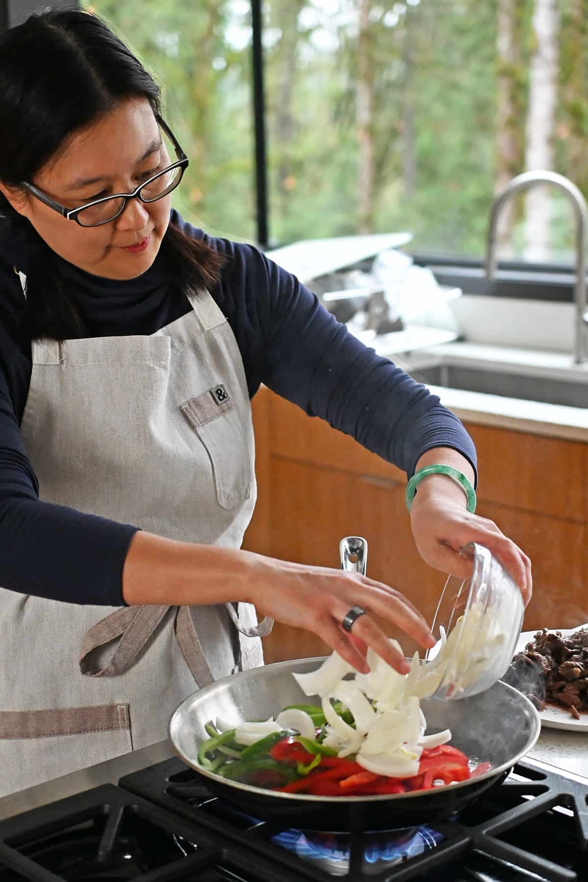 An Asian woman in glasses and braids is adding sliced onions to a hot skillet filled with sliced red and green bell peppers.
