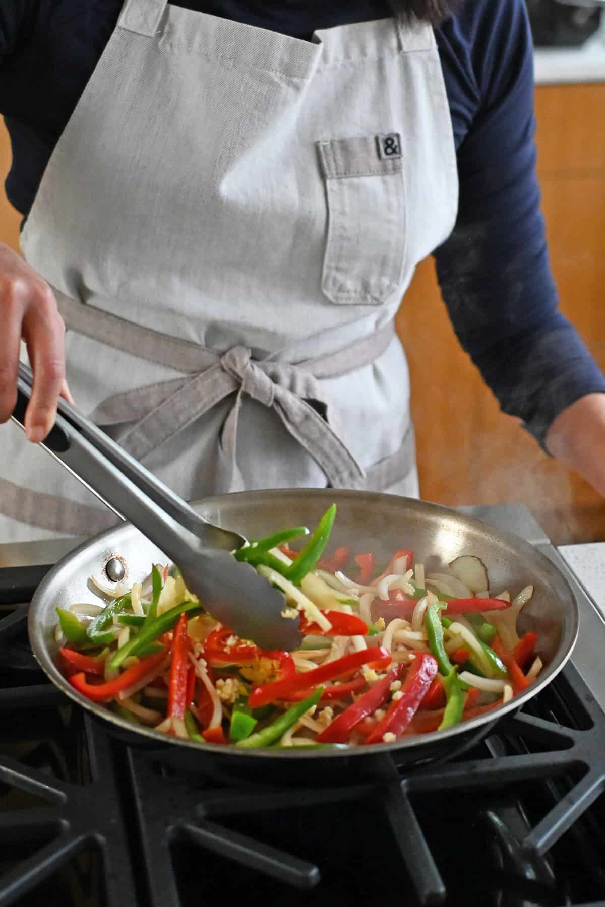 A pair of tongs is tossing sliced onions and belle peppers in a stainless steel skillet.