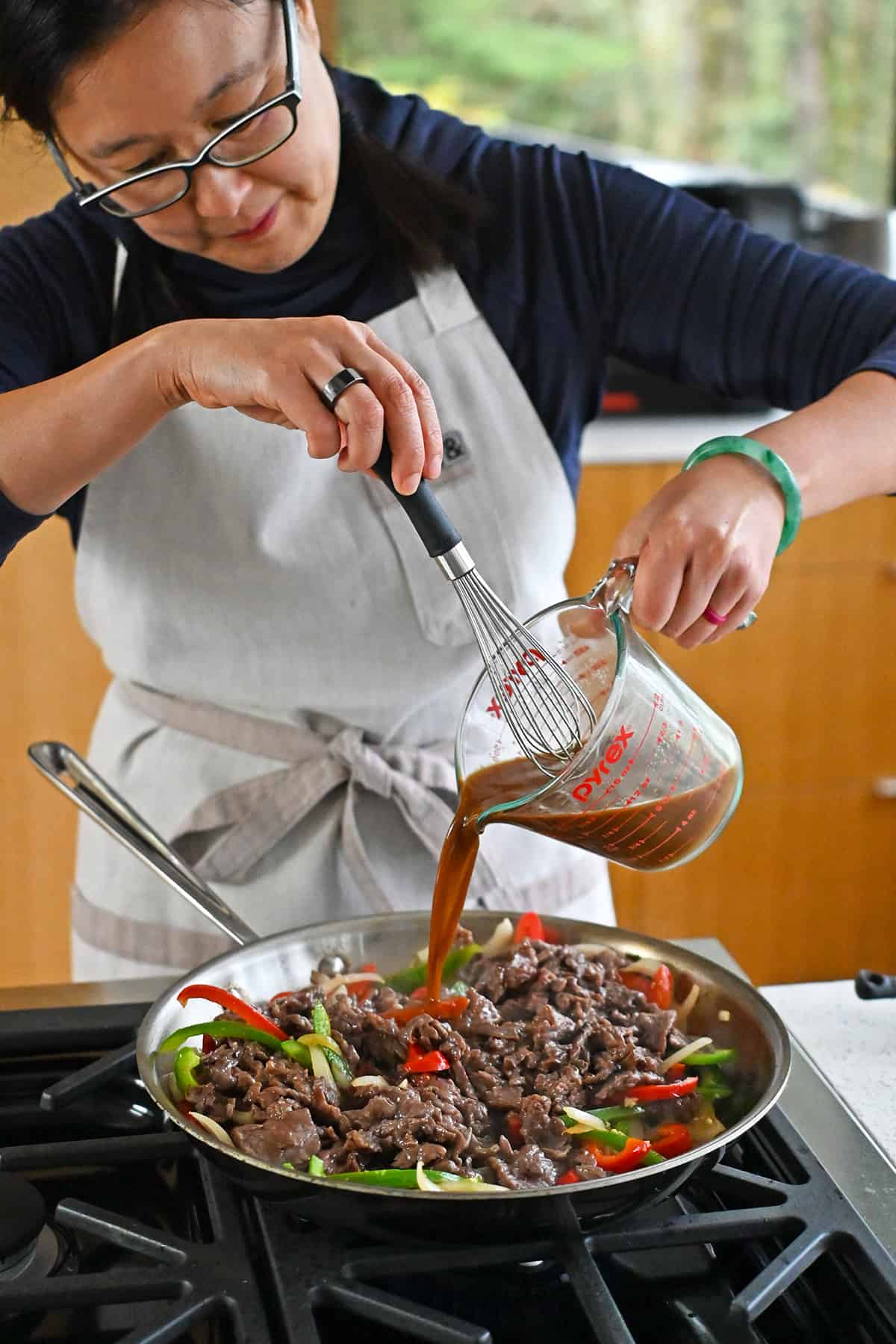 Pouring a brown sauce into a pan filled with cooked flank steak, sliced onions, and bell peppers.