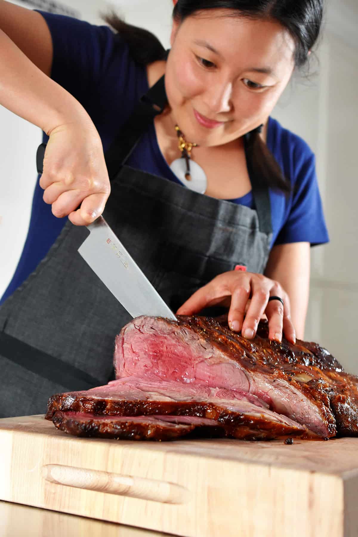 A smiling Asian woman slicing a medium rare prime rib roast into thin slices on a wooden cutting board.