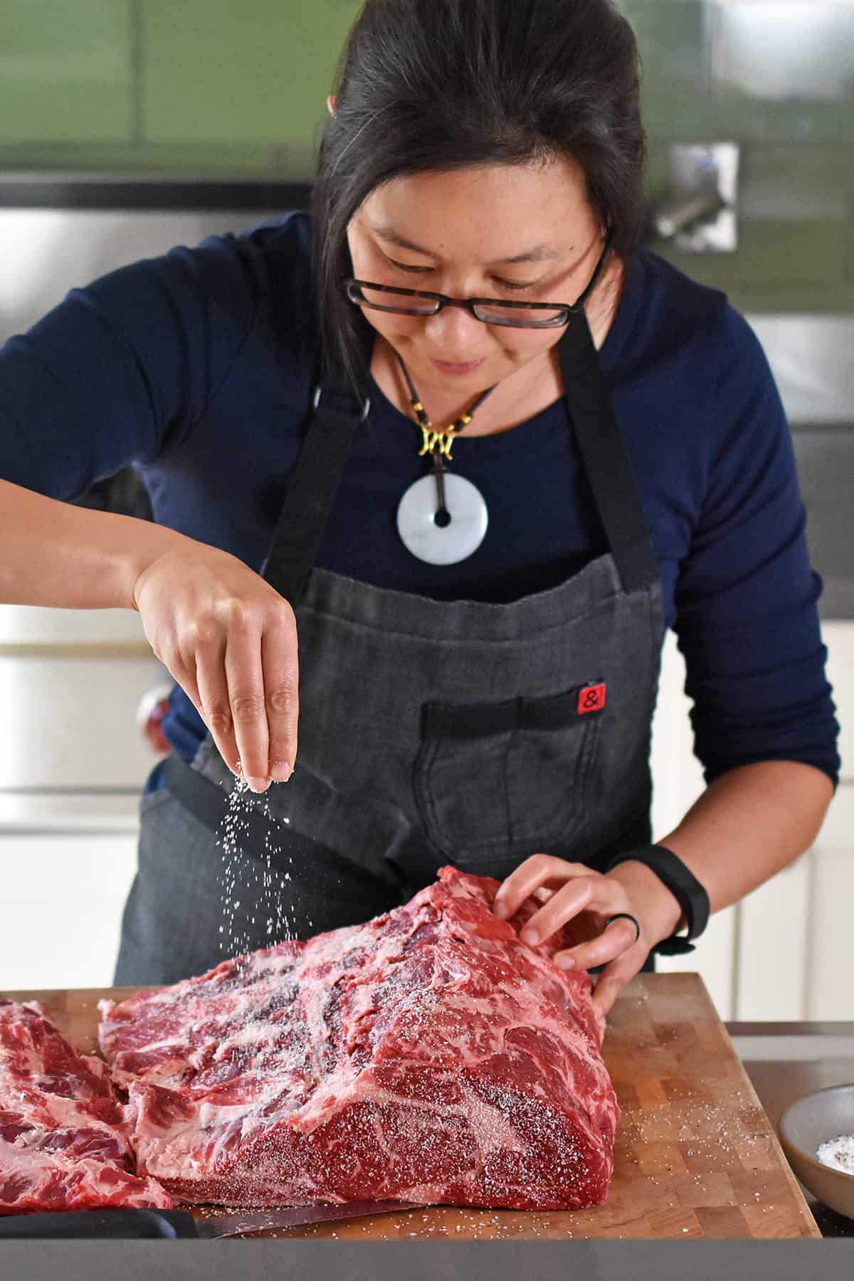 An Asian woman in glasses is sprinkling salt and pepper on a boneless prime rib roast.
