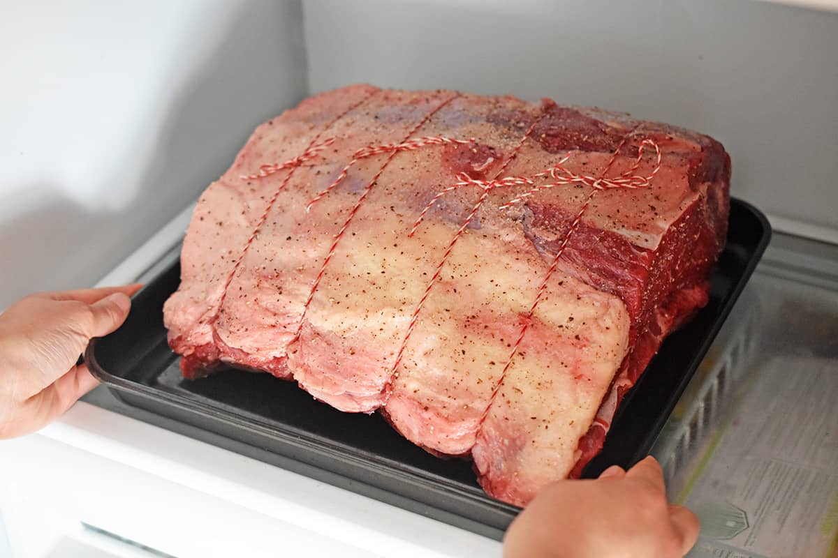 A seasoned and raw prime rib roast is being placed in the refrigerator, uncovered.