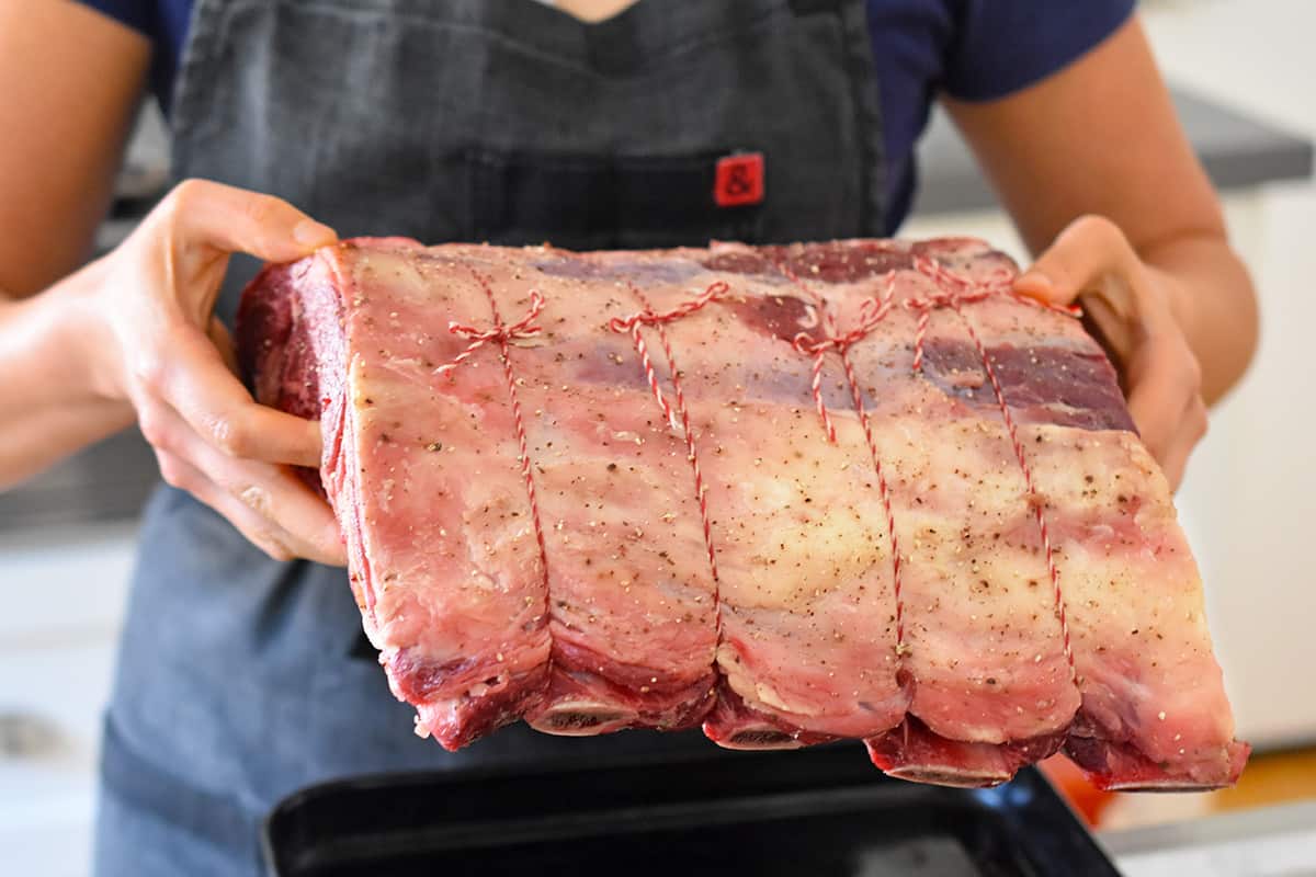 Someone placing a raw seasoned and tied prime rib roast into a small rimmed baking sheet.
