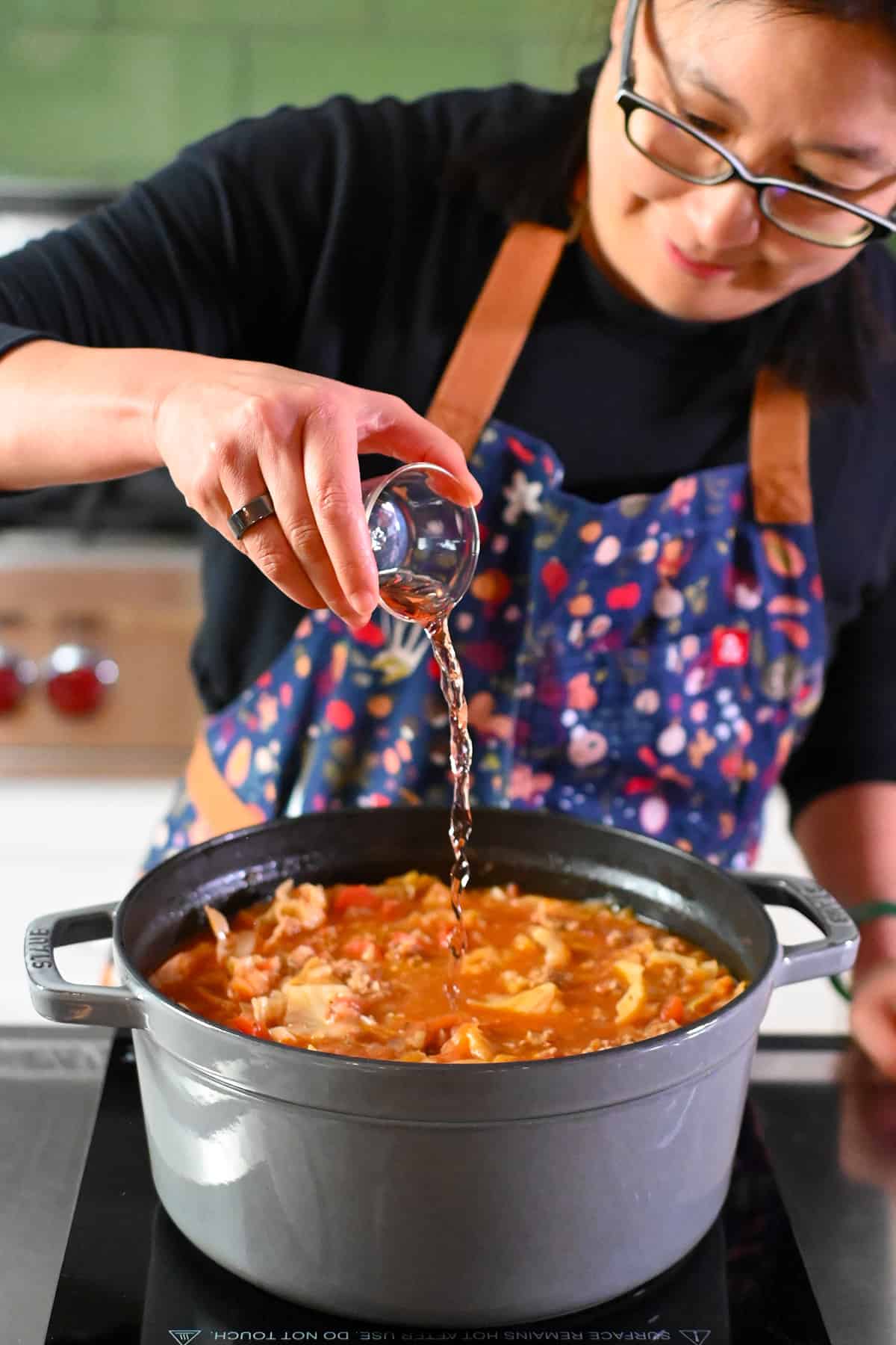 An Asian woman in glasses is adding some red wine vinegar to a gray stockpot filled with cabbage roll soup.