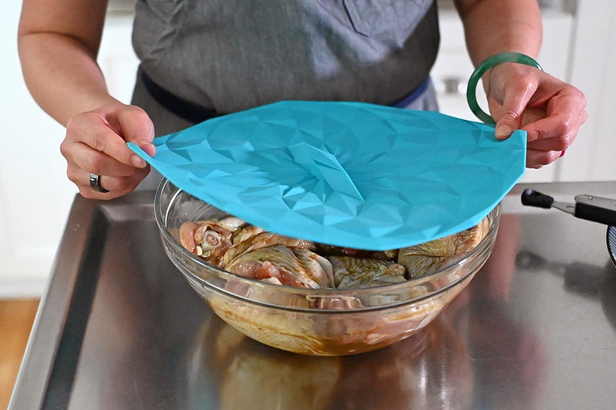 Covering a glass bowl with marinated raw chicken thighs with a blue silicone lid.