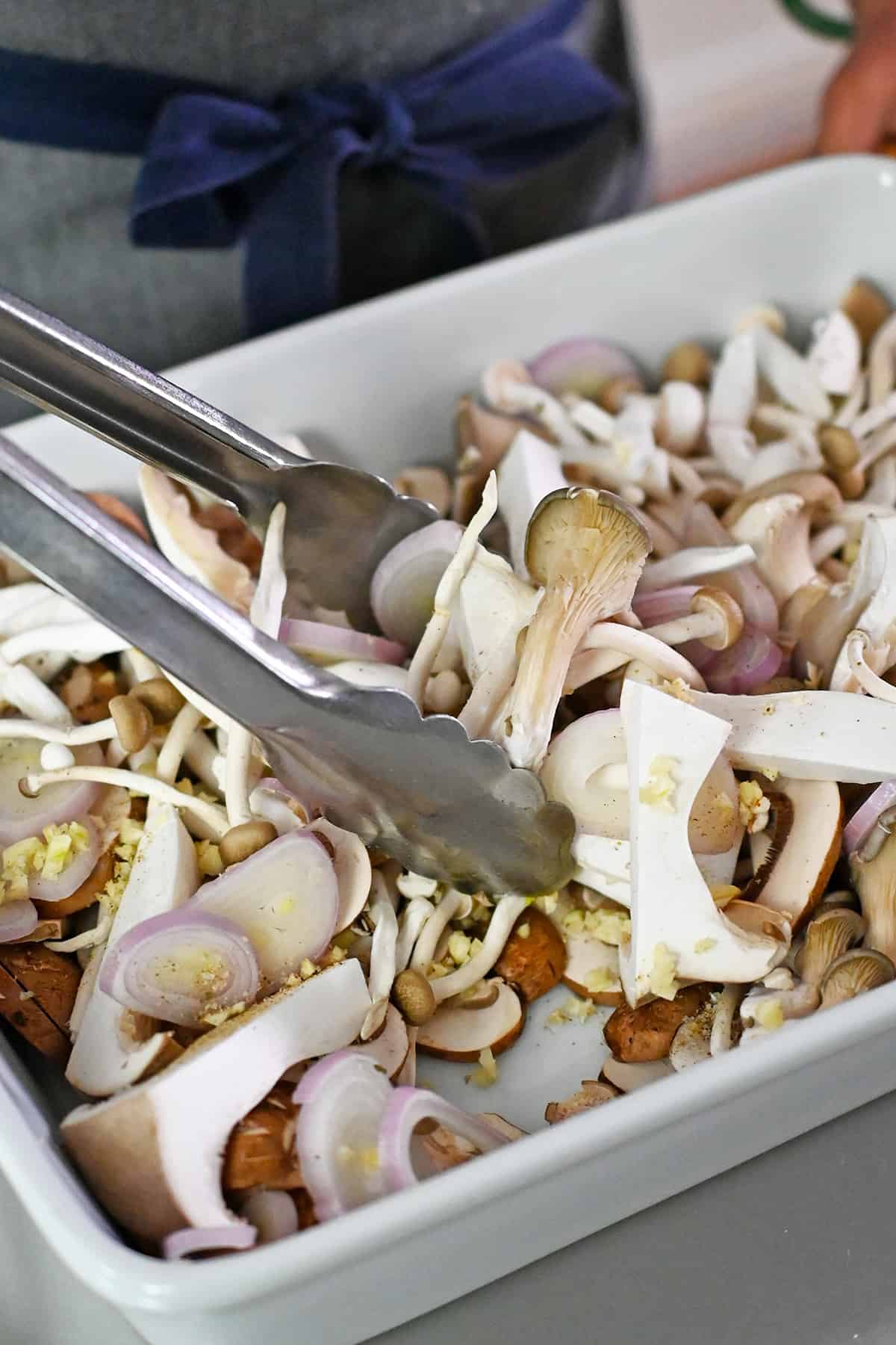 Tossing raw mushrooms, sliced shallots, minced garlic, and seasoning in a white casserole dish with a pair of tongs.