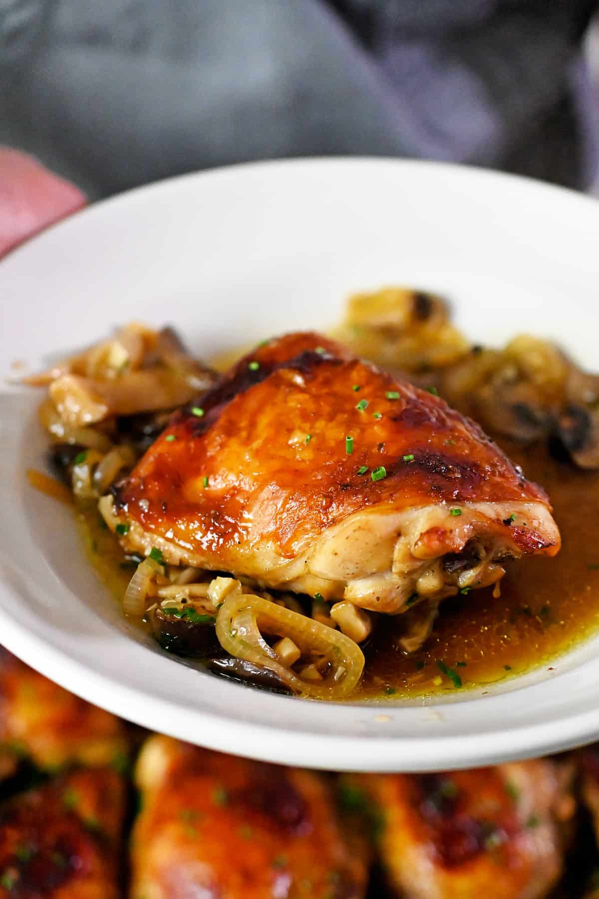 A white shallow dish topped with a golden brown chicken thigh, saucy mushrooms, and tender shallots.