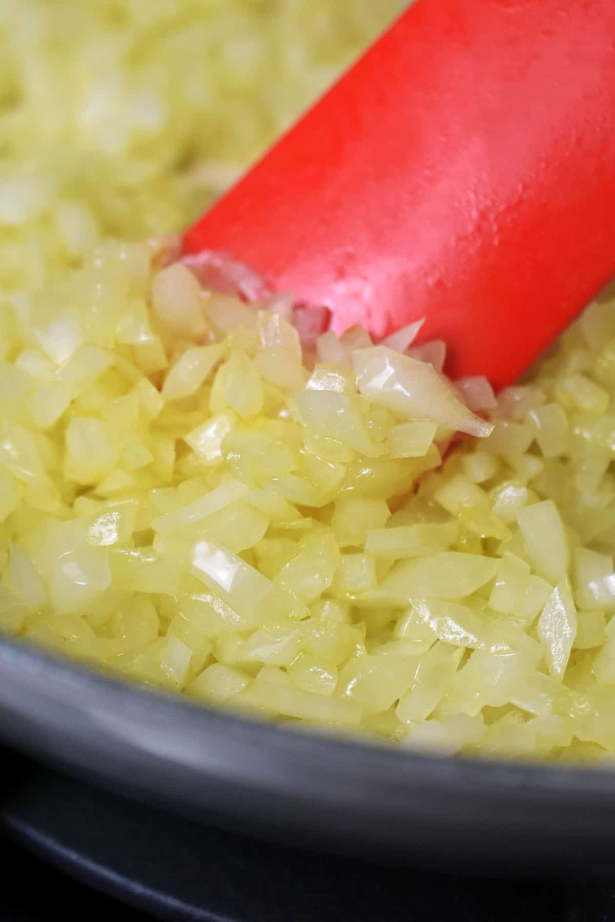 A red silicone spatula is pushing some softened sautéed chopped onions in a gray enamel cast iron skillet.