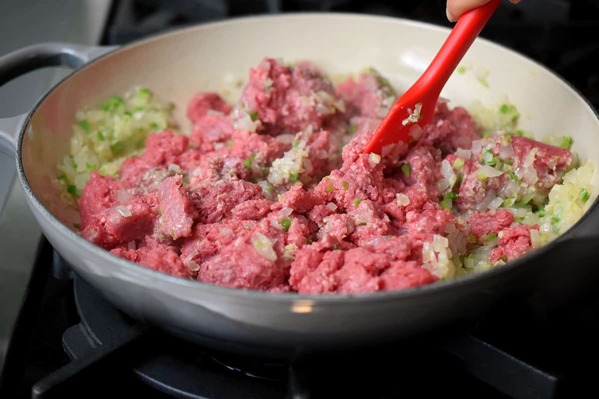 Adding raw ground meat to a skillet filled with sautéed chopped onions, minced garlic, minced ginger, and chopped jalapeños to make the filling for an Indian-spiced shepherd's pie.