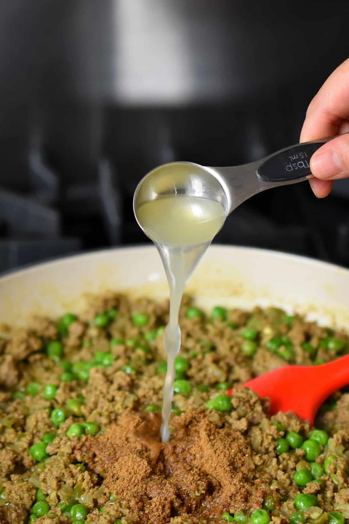 Adding a spoonful of lemon juice and ground Indian spices to a skillet filled with cooked ground beef, onions, and green peas.