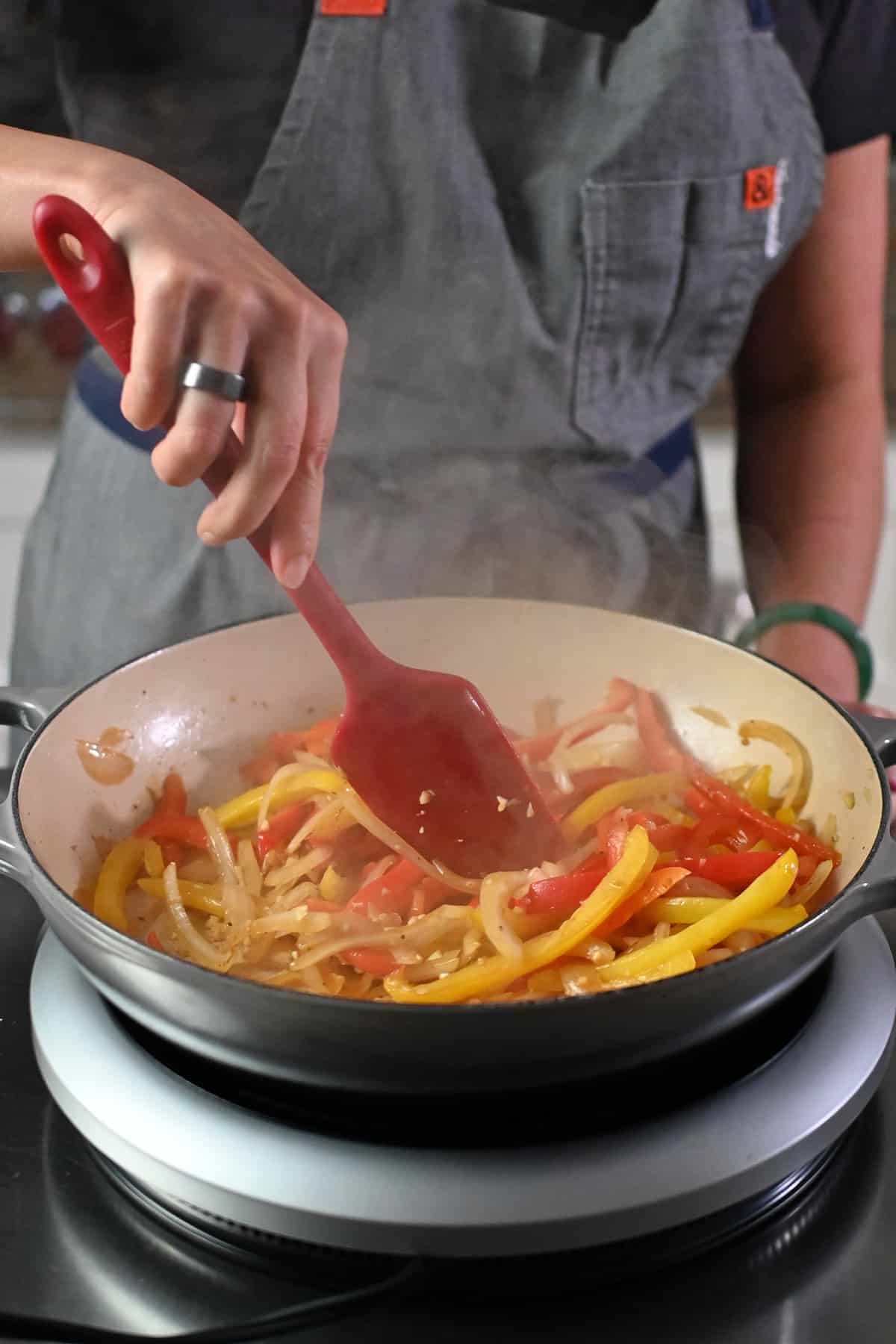 A red silicone spatula is stirring thinly sliced bell peppers and onions in a gray cast iron skillet to make shrimp fajitas.
