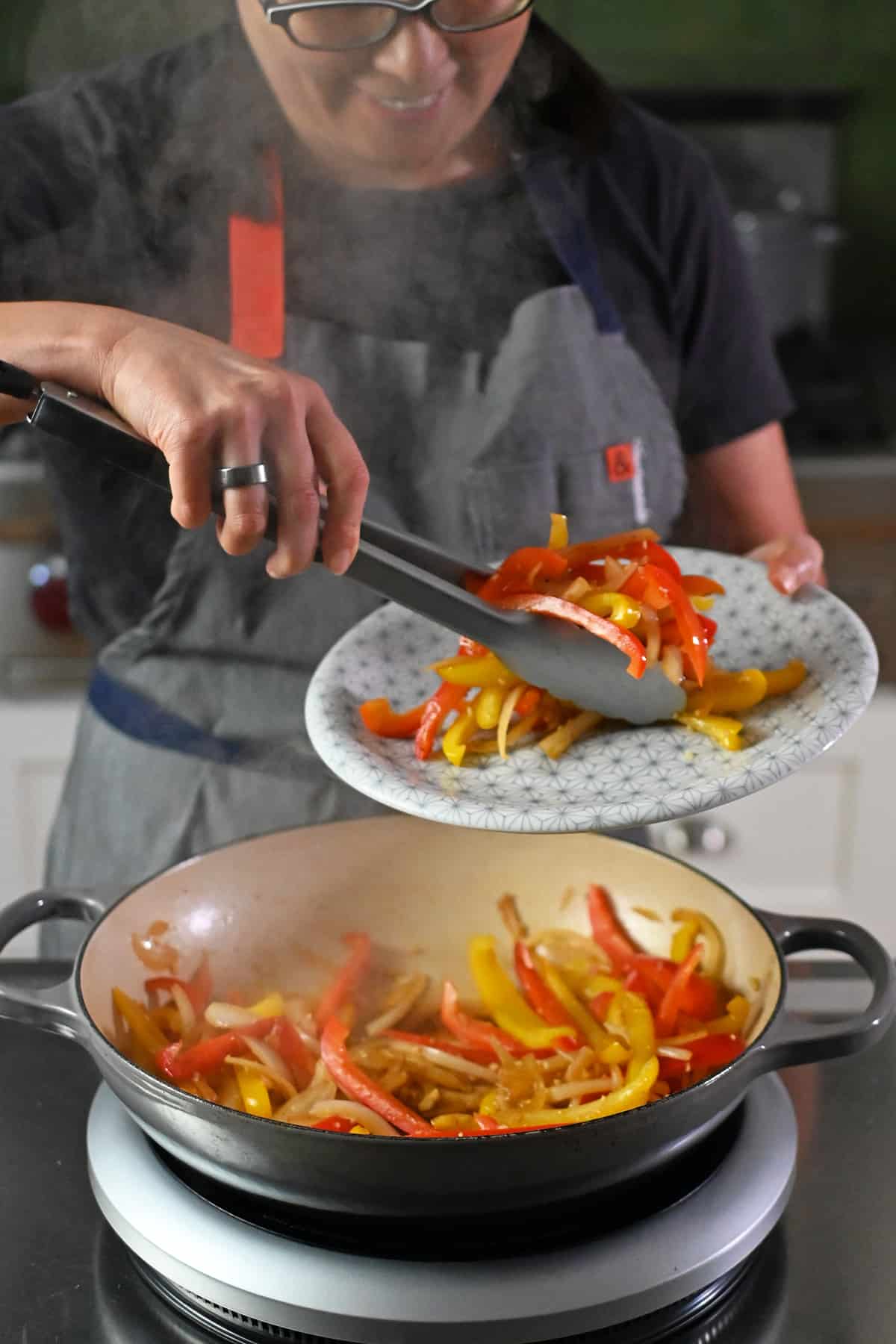 A smiling Asian woman with glasses is transferring cooked fajita vegetables from a cast iron pan to a platter with a pair of tongs.