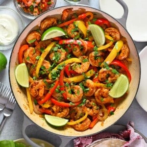 An overhead shot of a skillet filled with shrimp fajitas surrounded by bowls filled with guacamole, sour cream, and salsa, fresh lettuce leaves, and tortillas.
