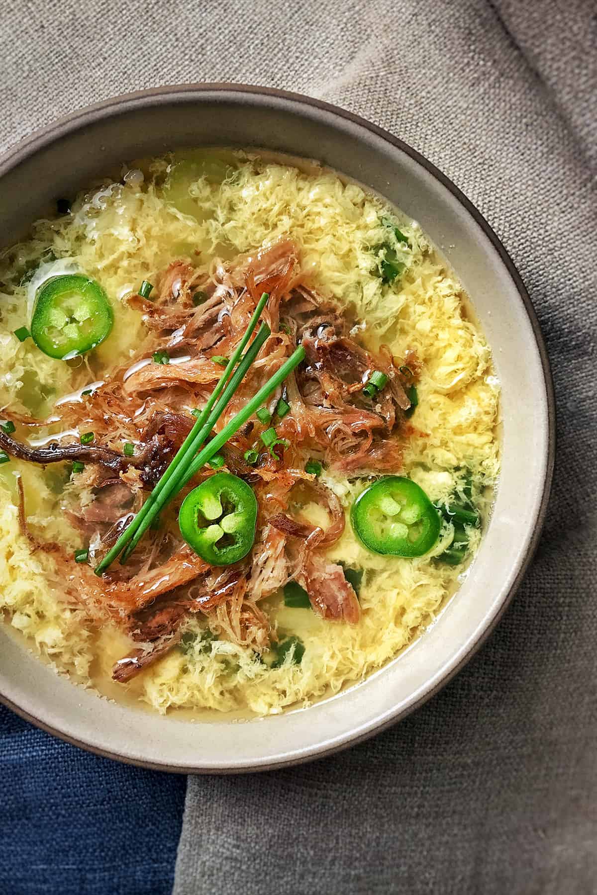 A bowl of egg drop soup topped with Kalua Pig, chives, and peppers.