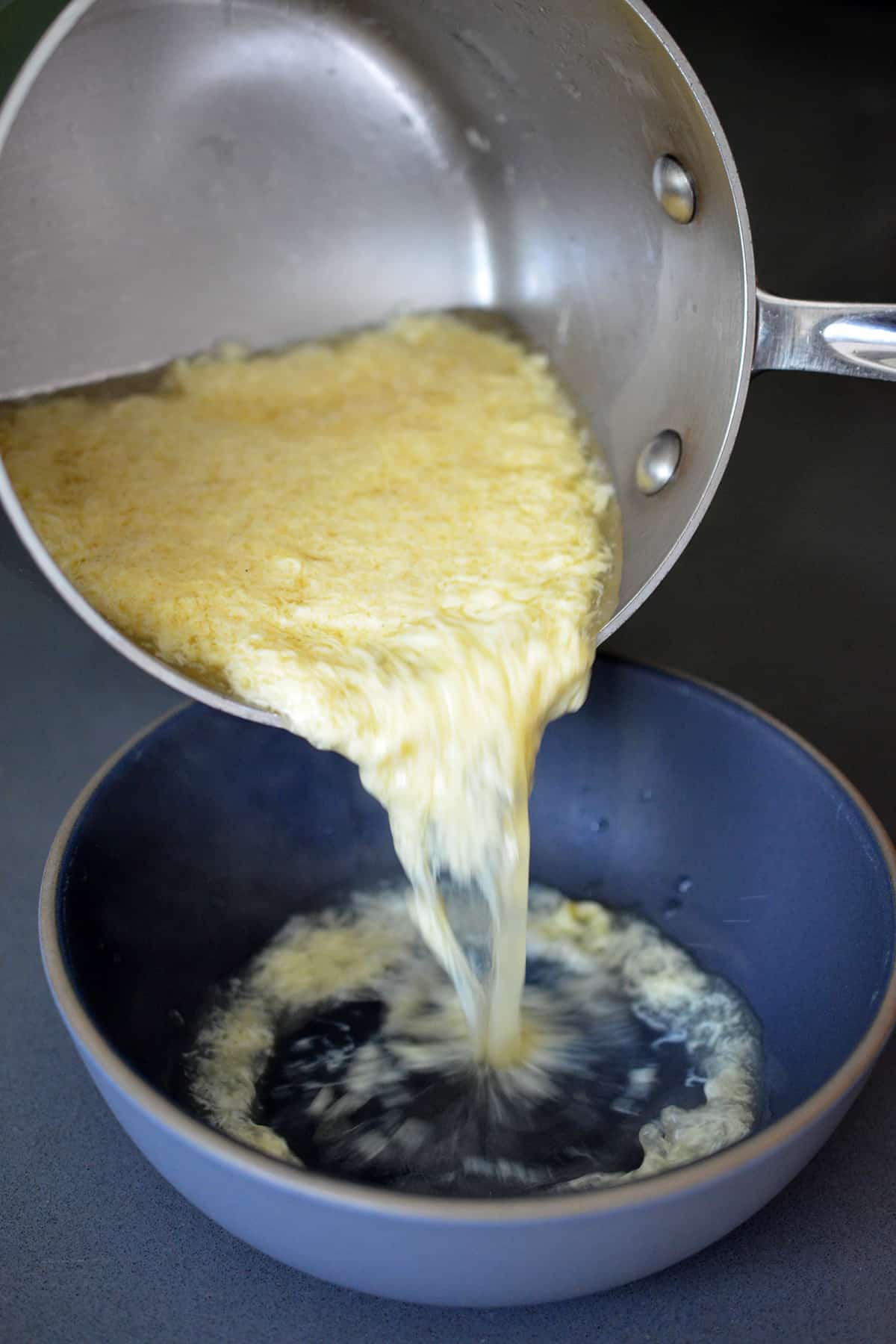 Pouring egg drop soup from a small saucepan into a blue bowl.