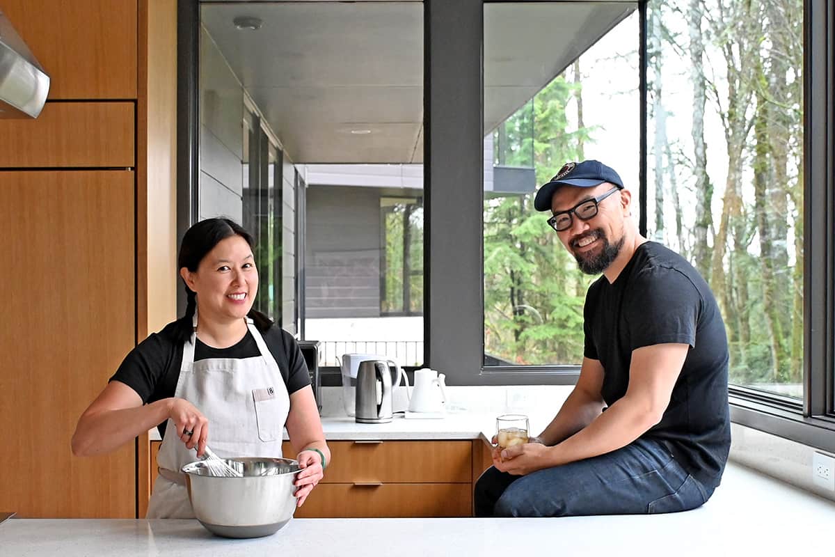 Photograph of a smiling Asian couple in an airy, modern kitchen; the woman is in an apron and stirring ingredients in a mixing bowl while the man is sitting on the countertop holding a drink.