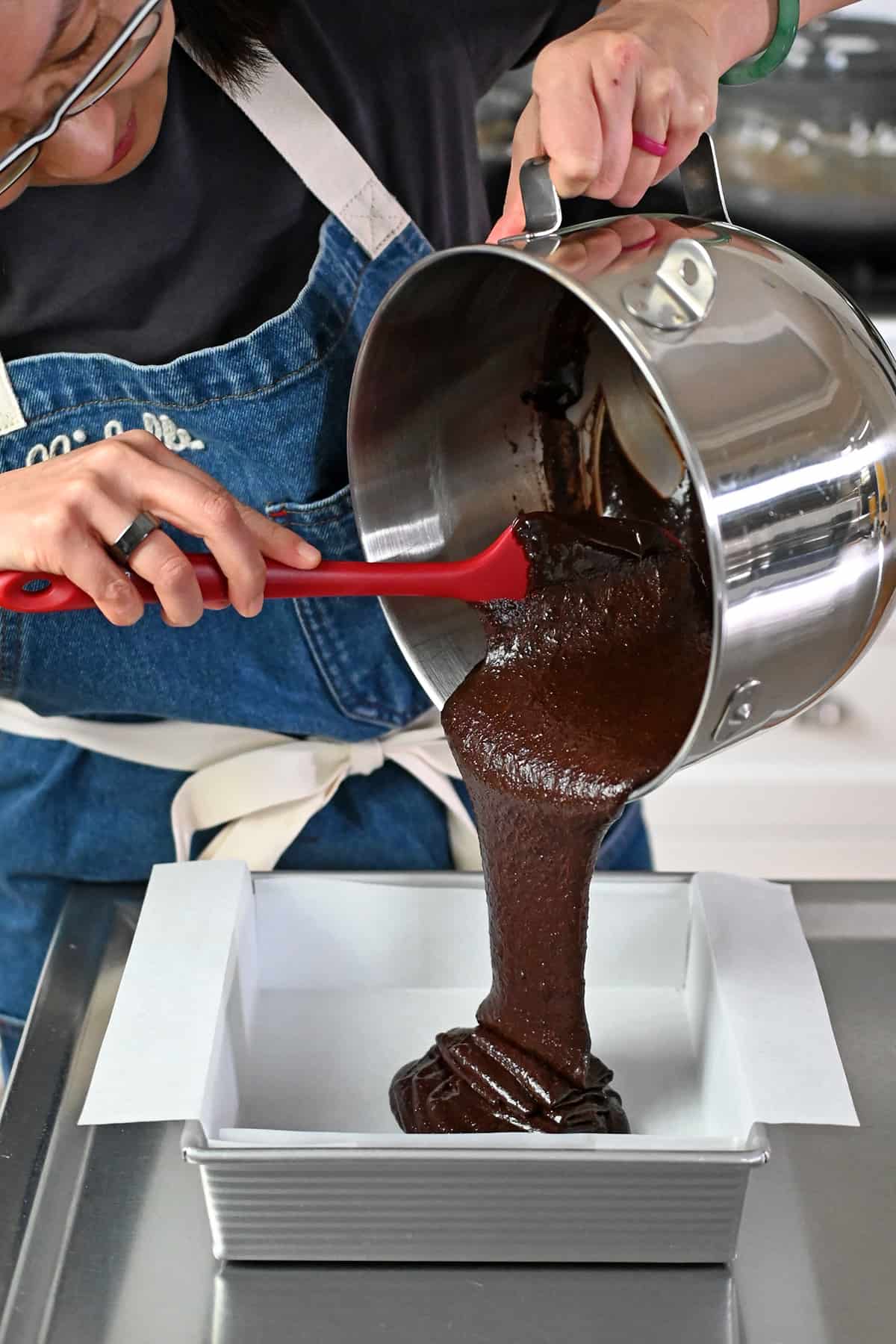 An Asian woman in glasses is pouring paleo brownie batter into a parchment lined square baking pan made out of aluminum.