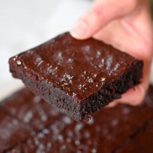 A hand is holding a square of paleo brownies above a cutting board filled with the remaining paleo brownies.