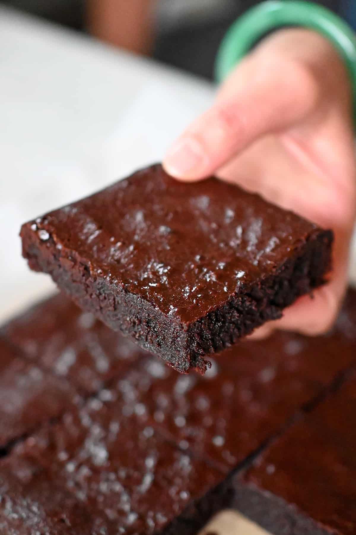 A hand is holding a square of paleo brownies above a cutting board filled with the remaining paleo brownies.