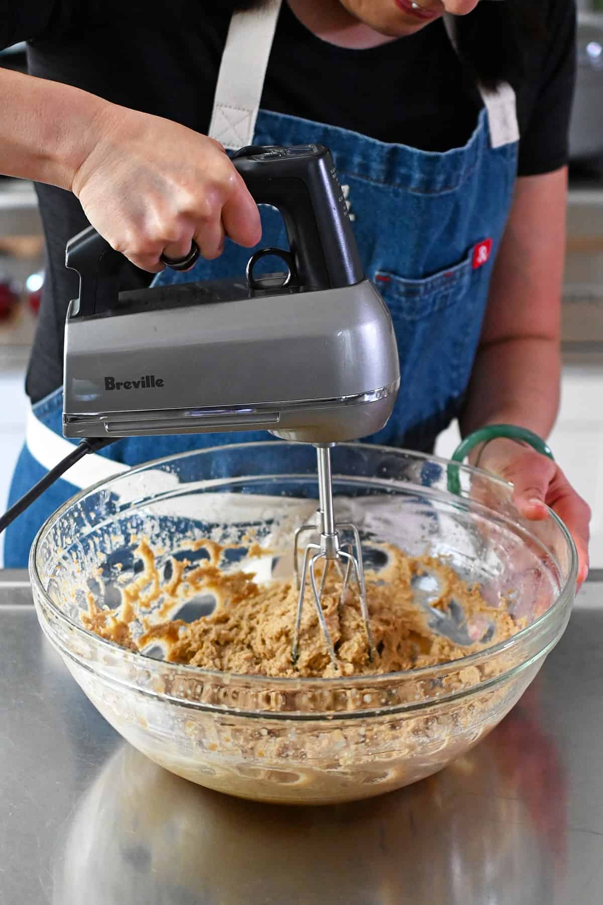 A person in a blue apron is using a hand mixer to combine the batter for paleo banana cookies in a glass mixing bowl.