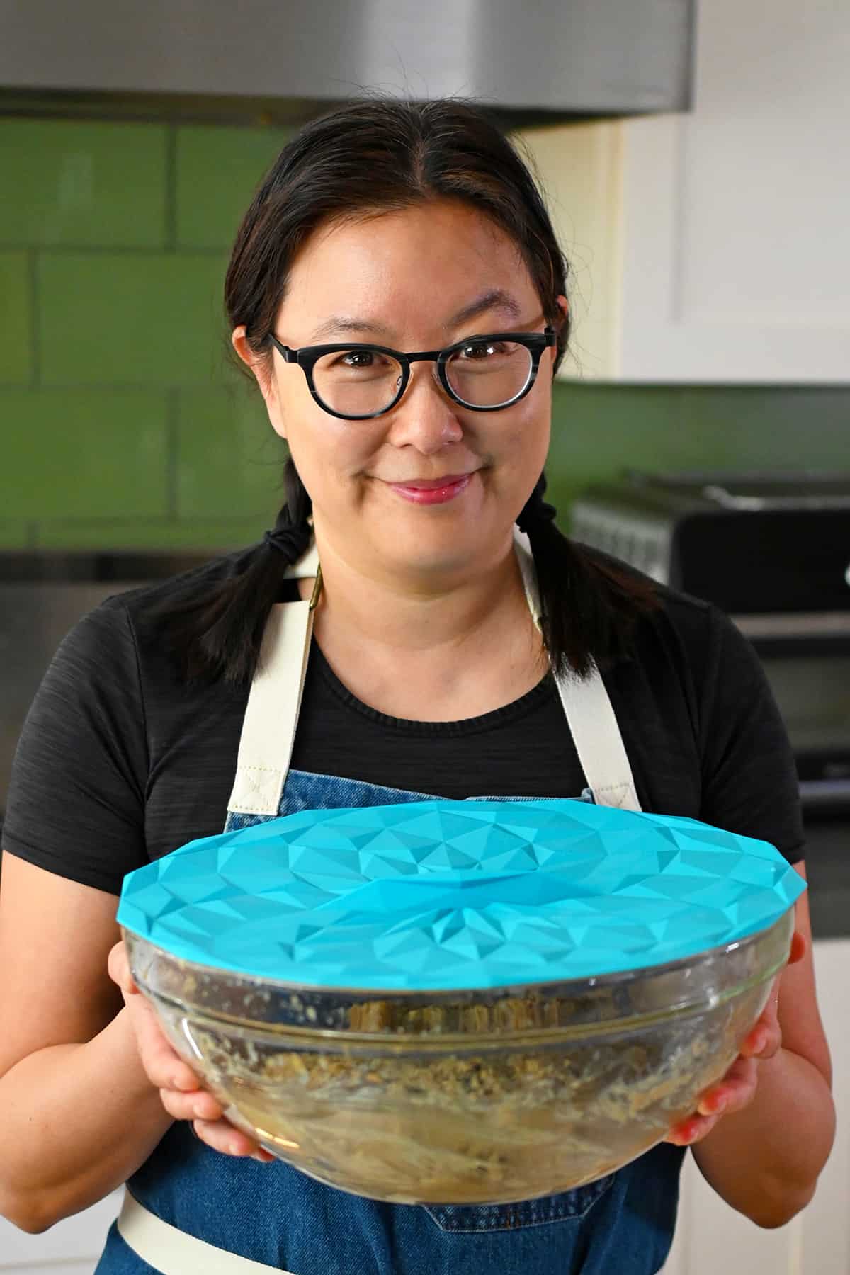 A smiling Asian woman wearing glasses holds a glass bowl full of paleo banana cookie batter covered with a blue silicone lid.