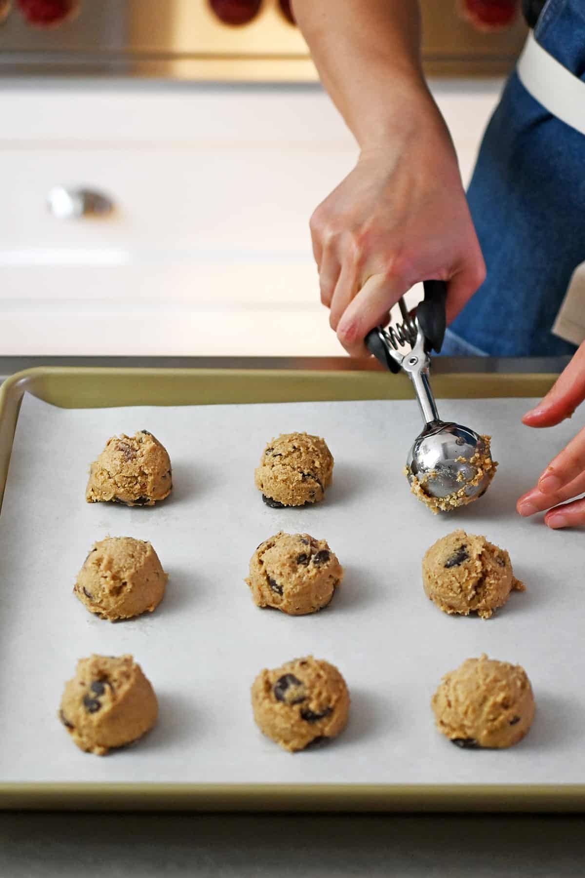 A rimmed baking sheet lined with parchment paper is covered with balls of raw banana cookie dough that are portioned with a medium ice cream scoop.