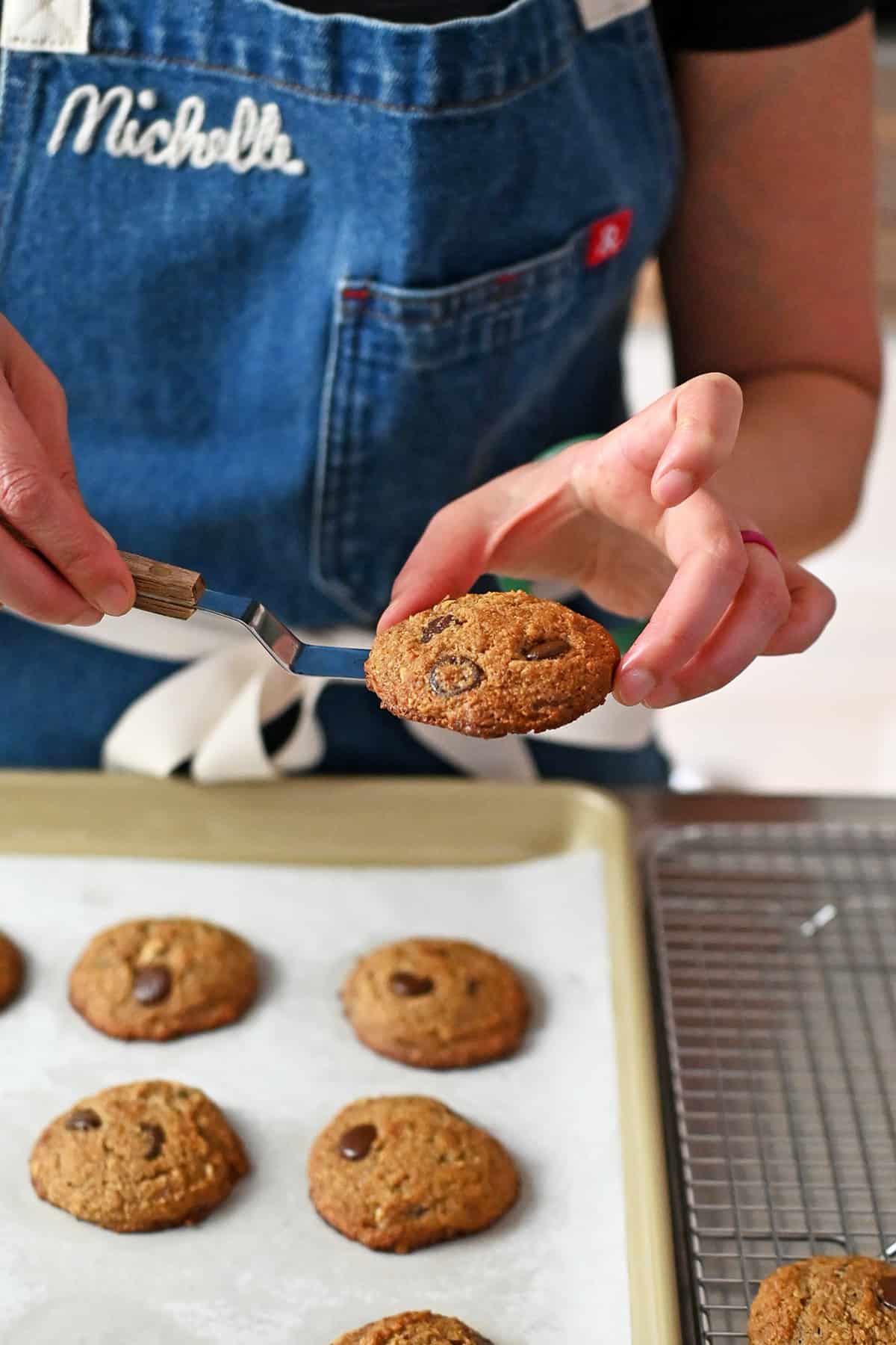 A person in a blue apron is using a small offset spatula to transfer freshly baked paleo banana cookies from a baking pan to a wire cooling rack,