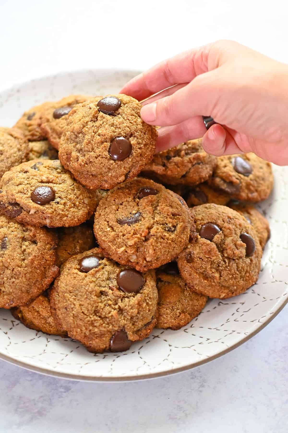 A hand is grabbing a paleo banana chocolate chip cookie from a white plate piled high with cookies.