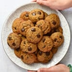 An overhead shot of two hands holding a white plate piled high with paleo banana chocolate chip cookies.