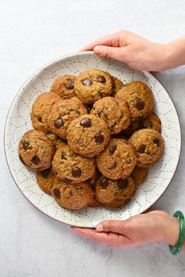 An overhead shot of two hands holding a white plate piled high with paleo banana chocolate chip cookies.