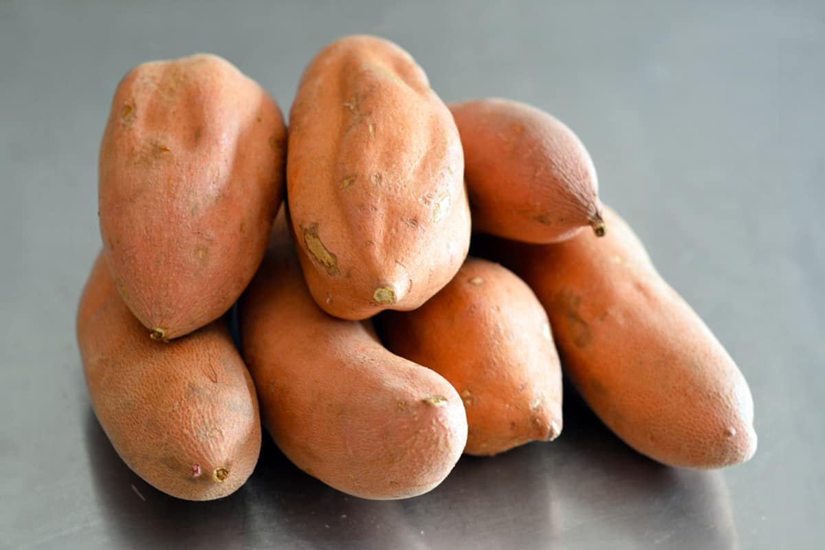 A pile of garnet yams or red fleshed sweet potatoes on a stainless steel counter.
