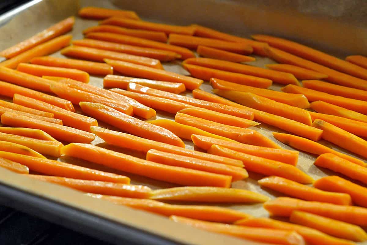 Sweet potato fries are in a single layer in a parchment lined rimmed baking sheet about to be baked in the oven.