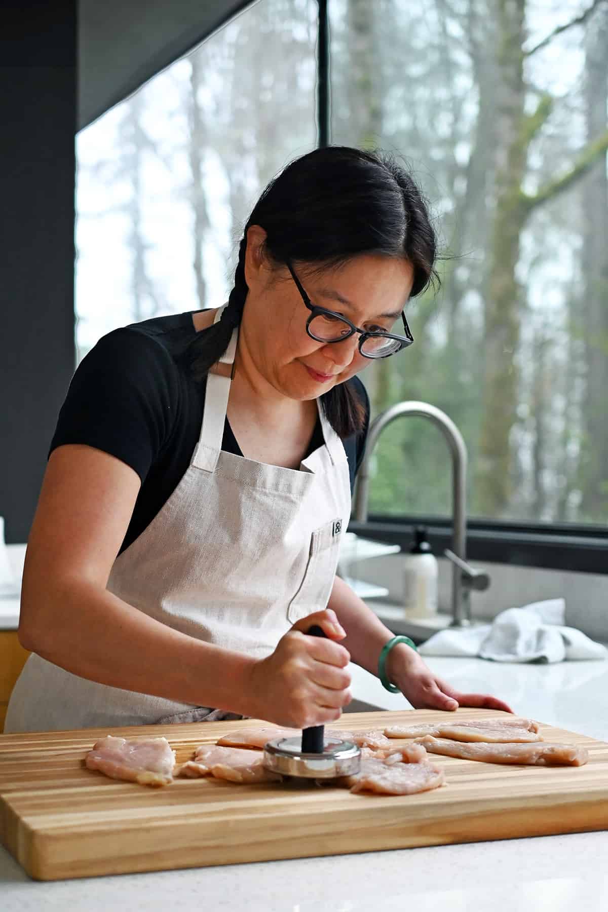 An Asian woman is using a meat pounder to flatten some raw chicken cutlets on a wooden cutting board before making pesto chicken.