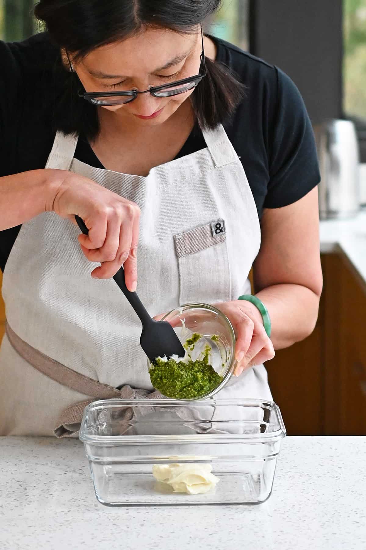 An Asian woman is adding fresh pesto to a glass container with some mayonnaise inside.