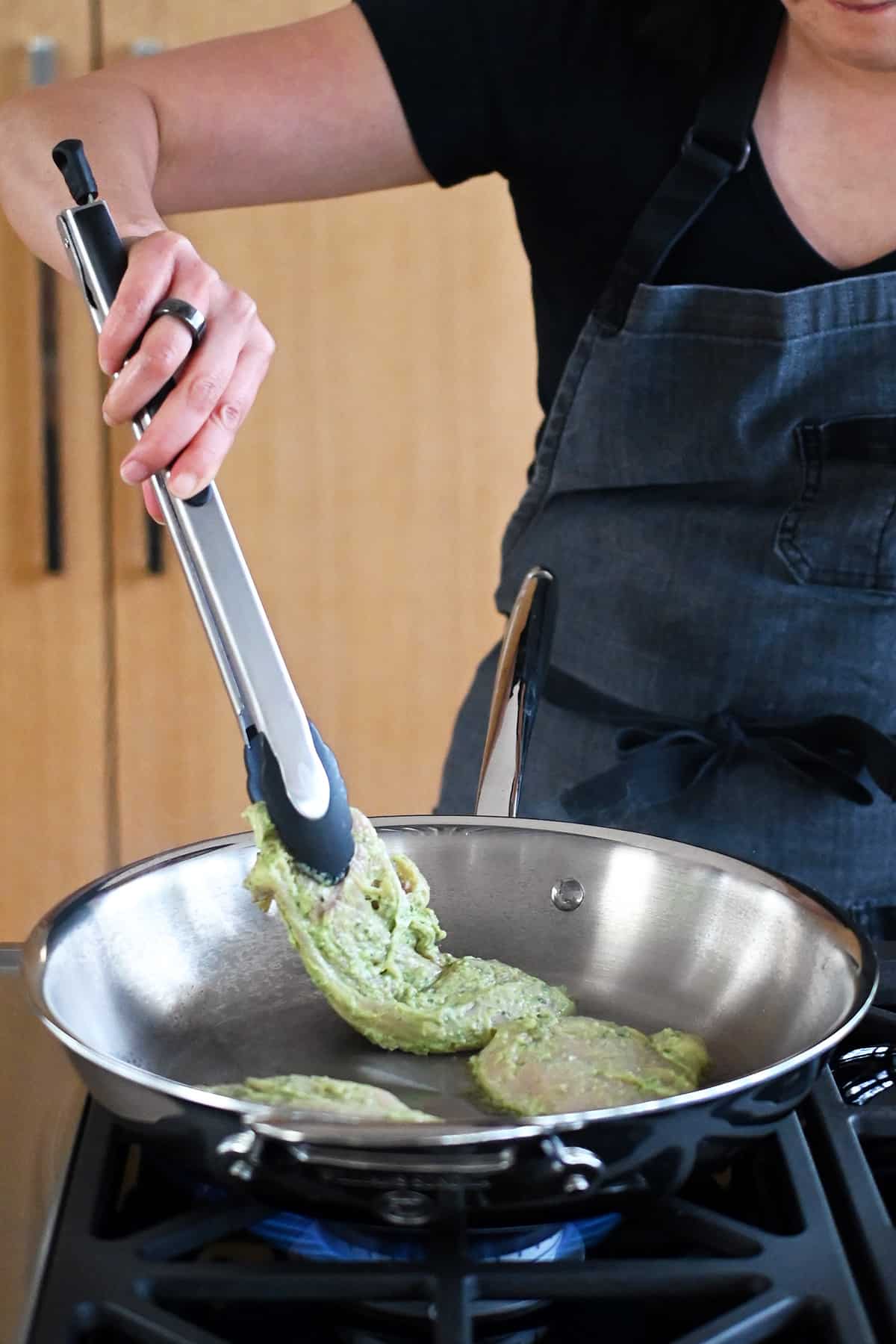 Adding pesto chicken to a stainless steel skillet with a pair of tongs.