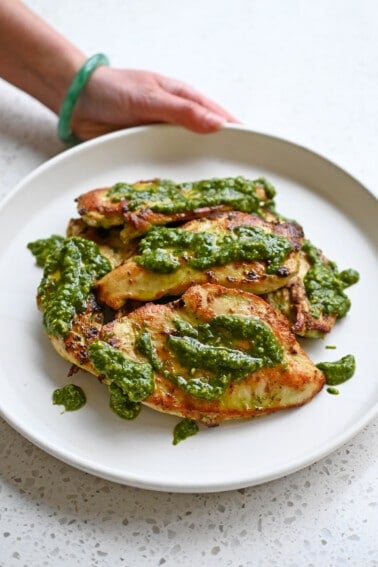A hand is holding a white plate filled with pesto chicken cutlets topped with extra pesto.