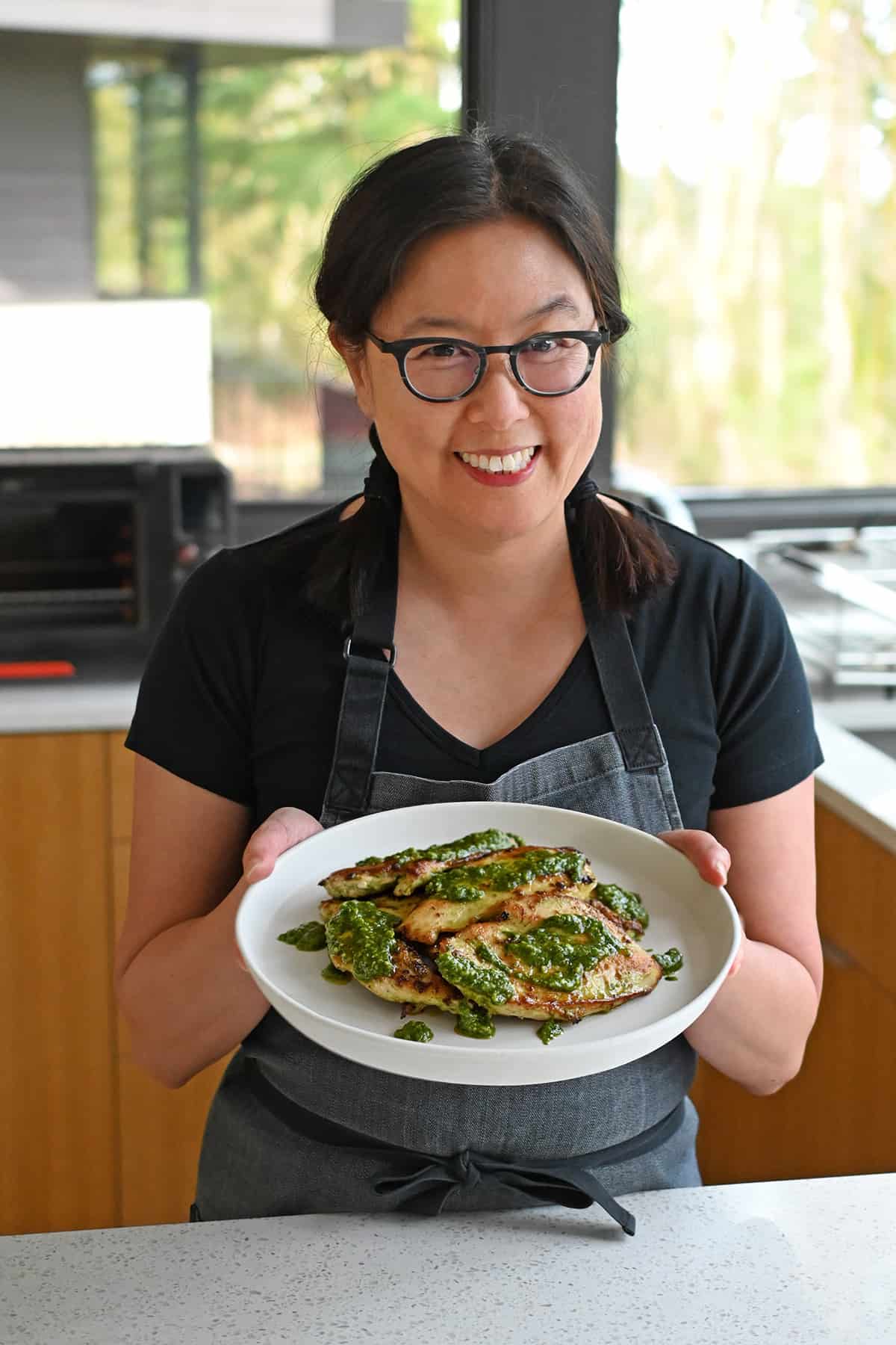 A smiling Asian woman wearing glasses is holding a plate filled with pesto chicken in a sunny kitchen.