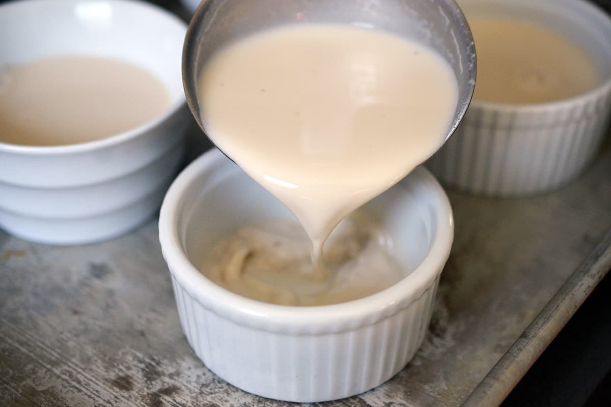 A ladle is pouring vanilla Panna Cotta into a white ramekin.